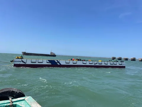 A flat-deck barge on the water with a jetty and vessel in the distance
